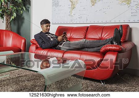 Young millennial businessman using his smart phone in the lobby of a workplace while laying on a couch; Sherwood Park, Alberta, Canada View Large Photo Image Stock Photograph - Young millennial businessman using his smart phone in the lobby of a workplace while laying on a couch; Sherwood Park, Alberta, Canada. Fotosearch