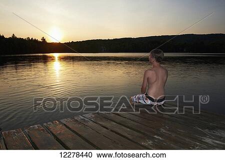 Stock Image - Boy sitting on the dock with feet in water at sunset; Lac des Neiges, Quebec, Canada. Fotosearch