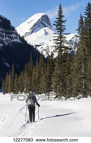 Stock Image - Female snowshoeing along a snow covered trail with snow covered mountain in the background and blue sky; Banff, Alberta, Canada. Fotosearch