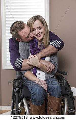 Stock Image - Husband embracing his disabled wife in their home; Spruce Grove, Alberta, Canada. Fotosearch