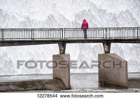 Water cascading down a reservoir overflow outlet with person in a pink coat watching from a bridge; Cumbria, England View Large Photo Image Picture - Water cascading down a reservoir overflow outlet with person in a pink coat watching from a bridge; Cumbria, England. Fotosearch