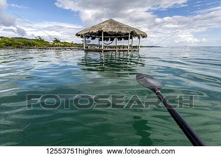 Stock Photograph - A boat paddle over turquoise water with a shelter at the end of the dock and the coastline of a Caribbean island; Honduras. Fotosearch