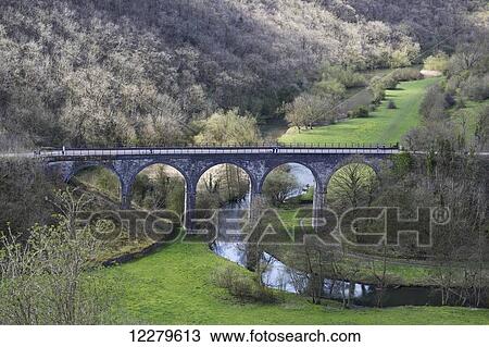 Stock Image - A bridge with arches crossing a small river in a valley; Derbyshire, England. Fotosearch