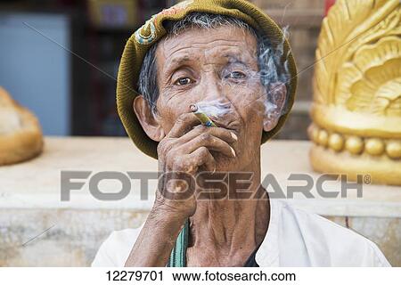 Stock Image - A grizzled older man smokes a cheroot while looking through the smoke; Myanmar. Fotosearch