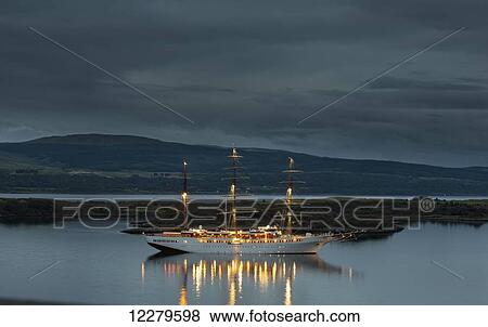 Stock Photo - A large white boat with masts illuminated on the tranquil water along the coast at dusk; Tobermory, Isle of Mull, Argyll and Bute, Scotland. Fotosearch