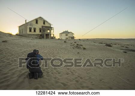 Picture - A person takes photos of the abandoned homes in the Namib desert at sunset; Kolmanskop, Namibia. Fotosearch