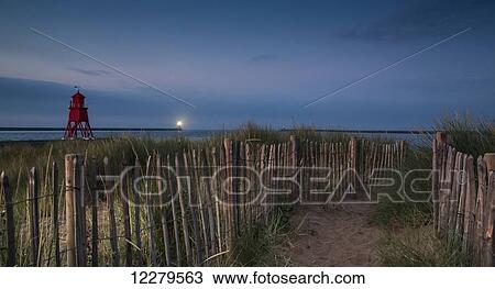 Stock Image - A sand pathway lined with a wooden fence leading to the beach with the Herd Groyne Lighthouse on the water's edge; South Shields, Tyne and Wear, England. Fotosearch