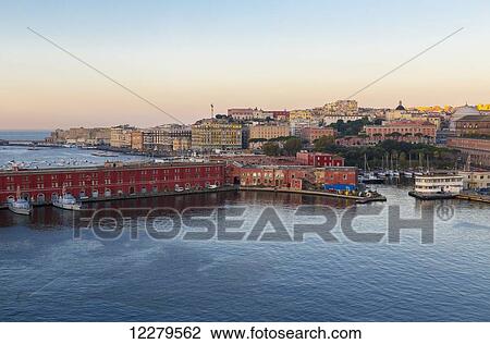 Stock Image - A wide landscape view of the port of Naples, home of Italy's navy seen in the foreground with the historical ancient town of Naples in the background at dawn; Naples, Campania, Italy. Fotosearch