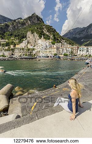 Stock Photograph - A young female tourist poses for a photo on the boardwalk with the city of Amalfi in the background on a vacation in Italy; Amalfi, Province of Salerno, Italy. Fotosearch
