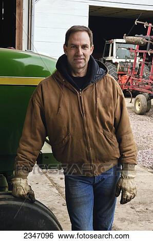 Stock Photograph - Agriculture - A farmer stands next to his John Deere tractor prior to spring cultivating and planting, with other farm machinery in the background / Iowa, USA.. Fotosearch