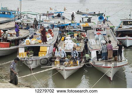 Stock Image - Boats tied up on the Mandalay shore of the Irrawaddy River teaming with activity as they load goods and passengers; Mandalay, Myanmar. Fotosearch