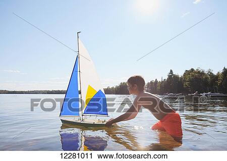 Stock Image - Boy standing in lake playing with sailboat; Ontario, Canada. Fotosearch