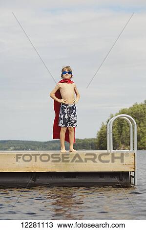 Stock Image - Boy standing on dock wearing goggles and towel pretending to be a super hero; Ontario, Canada. Fotosearch