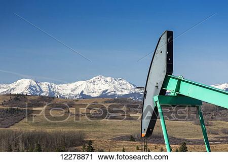 Stock Image - Close up of pump jack with rolling hills and snow covered mountains in the background with blue sky; Alberta, Canada. Fotosearch