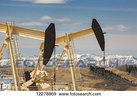 Close up of two pump jacks with rolling hills and snow covered mountains in the background with blue sky and clouds; Alberta, Canada View Large Photo Image Stock Photo - Close up of two pump jacks with rolling hills and snow covered mountains in the background with blue sky and clouds; Alberta, Canada. Fotosearch