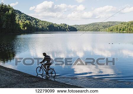 Stock Photo - Cyclist riding through shallow water at the edge of a lake; Waterbury, Vermont, United States of America. Fotosearch