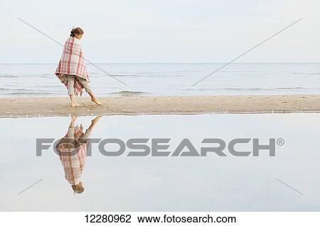 Stock Image - Girl reflected in water standing on beach wrapped in blanket; Toronto, Ontario, Canada. Fotosearch