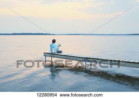 Picture - Girl with her dog sitting at end of dock at sunrise on Balsam Lake; Ontario, Canada. Fotosearch