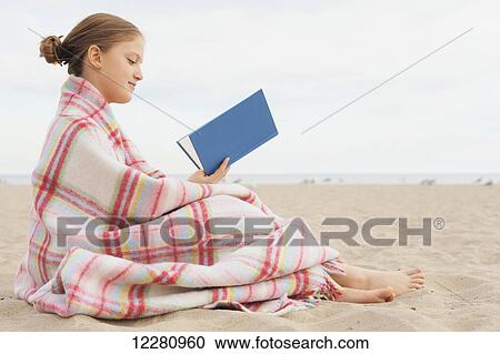 Stock Image - Girl wrapped in a blanket sitting on beach reading a book; Toronto, Ontario, Canada. Fotosearch