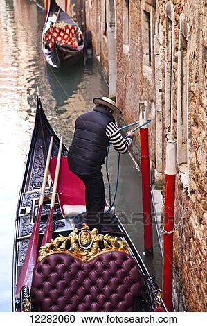 Stock Image - Gondolier tying his gondola to a post beside a building on a canal; Venice, Italy. Fotosearch