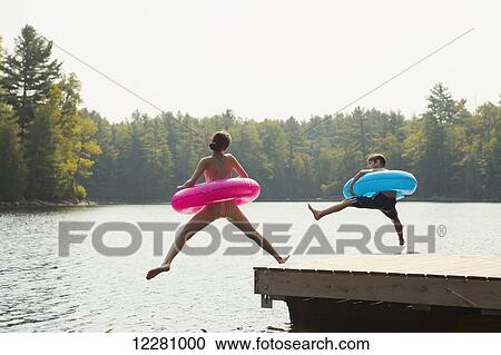 Stock Image - Kids jumping off end of dock holding inflatable rings on Crystal Lake; Ontario, Canada. Fotosearch