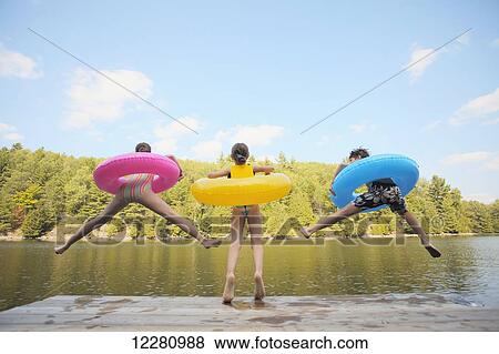Stock Photo - Kids jumping off end of dock holding inflatable rings on Crystal Lake; Ontario, Canada. Fotosearch