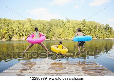 Stock Photo - Kids jumping off end of dock holding inflatable rings on Crystal Lake; Ontario, Canada. Fotosearch
