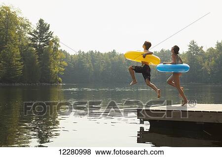 Stock Photo - Kids jumping off end of dock holding inflatable rings on Crystal Lake; Ontario, Canada. Fotosearch