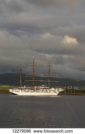 Stock Photograph - Large white boat with masts along the shoreline with storm clouds; Tobermory, Isle of Mull, Argyll and Bute, Scotland. Fotosearch
