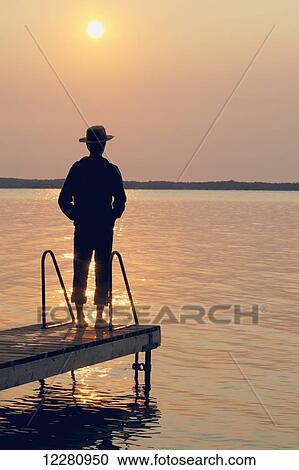 Stock Image - Man standing at end of dock at sunrise on Balsam Lake; Ontario, Canada. Fotosearch