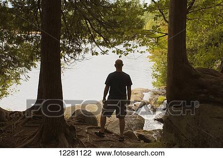 Stock Image - Man standing in forest looking out over lake; Bracebridge, Ontario, Canada. Fotosearch