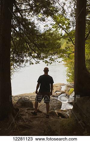 Stock Image - Man standing in forest looking out over lake; Bracebridge, Ontario, Canada. Fotosearch