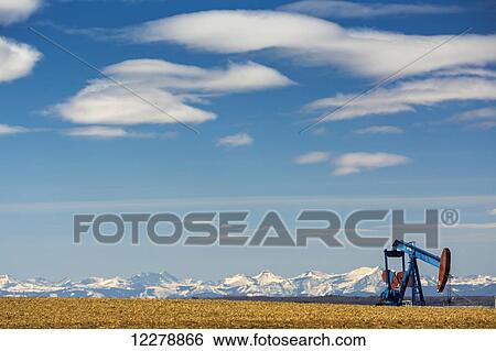 Stock Photograph - Pump jack in stubble field with snow covered mountains in the background with blue sky and clouds; Alberta, Canada. Fotosearch