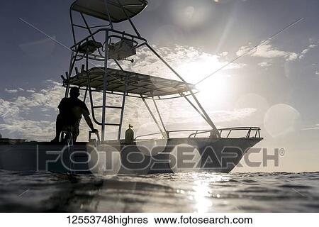Stock Photograph - Silhouette of a man on the deck of a boat on the water against a blue sky with sunlight; Bay Islands Department, Honduras. Fotosearch