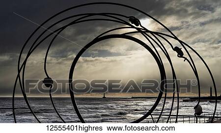 Stock Photograph - Silhouette of a sculpture and Roker Lighthouse in the distance along the shoreline of River Ware with storm clouds overhead; Sunderland, Tyne and Wear, England. Fotosearch