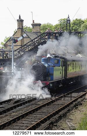 Stock Image - Steam from a passenger train traveling down the tracks as people watch from a walkway above the tracks; Cumbria, England. Fotosearch