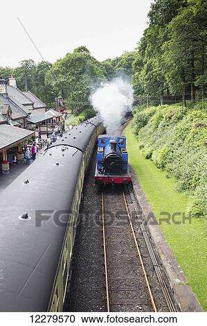 Stock Image - Steam from an engine traveling down the tracks as passengers wait to board a train at a station; Cumbria, England. Fotosearch