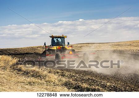 Stock Photo - Tractor tilling a hill side field with clouds and blue sky; Alberta, Canada. Fotosearch