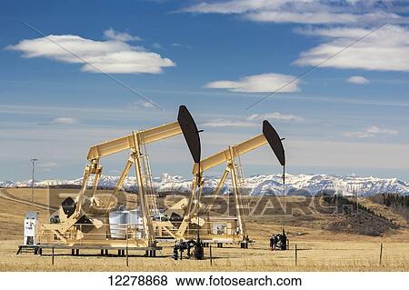 Two pump jacks on oil site with rolling hills and snow covered mountains in the background with blue sky and clouds; Alberta, Canada View Large Photo Image Stock Photo - Two pump jacks on oil site with rolling hills and snow covered mountains in the background with blue sky and clouds; Alberta, Canada. Fotosearch