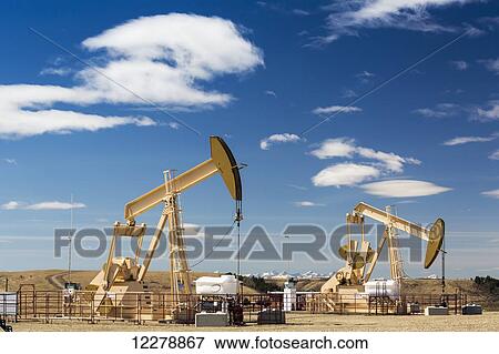 Stock Photo - Two pump jacks on oil site with snow covered mountains in the background and blue sky and clouds; Alberta, Canada. Fotosearch