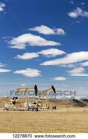 Stock Image - Two pump jacks with rolling hills and snow covered mountains in the background with blue sky and clouds; Alberta, Canada. Fotosearch