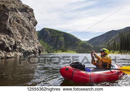 Stock Photograph - Woman photographing from her Packrafting on the Charley River in summertime, YukonCharley Rivers National Preserve; Alaska, United States of America. Fotosearch
