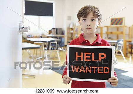 Stock Photo - Young boy standing in classroom holding help wanted sign; Toronto, Ontario, Canada. Fotosearch