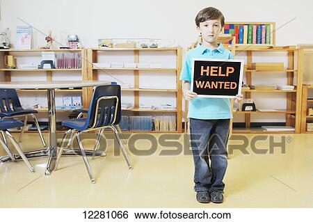 Stock Photograph - Young boy standing in classroom holding help wanted sign; Toronto, Ontario, Canada. Fotosearch