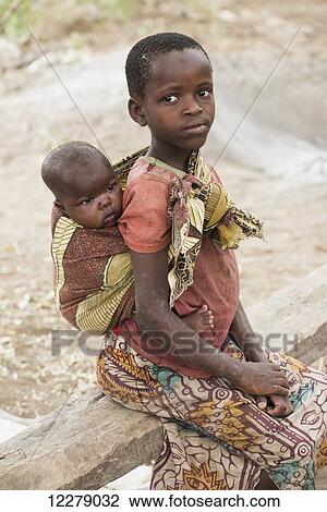 Stock Image - Young Datoga girl carries baby brother on back while sitting on a bench near Lake Eyasi; Tanzania. Fotosearch