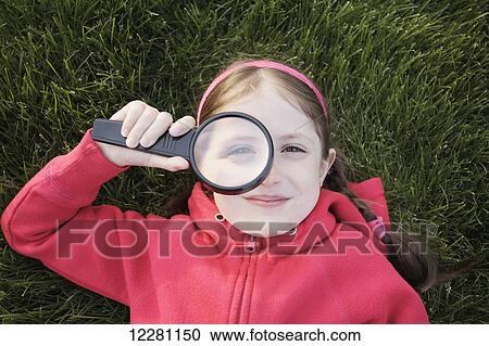 Stock Image - Young girl laying in grass holding a magnifying glass; Toronto, Ontario, Canada. Fotosearch