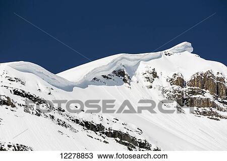 Stock Image - Close up of snow covered mountain peak with large overhanging cornice and deep blue sky; Alberta, Canada. Fotosearch