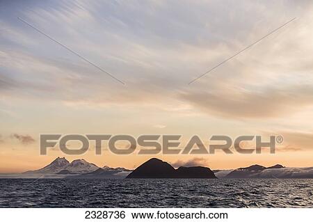 Stock Photograph - Isanotski Peaks (Ragged Jack) And Round Top Mountain On Unimak Island With Sankin Island In The Foreground After Sunset; Southwest Alaska, United States Of America. Fotosearch