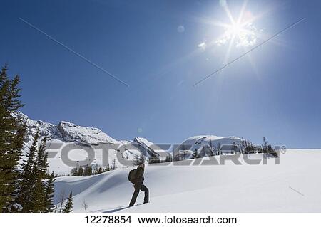 Male snowshoer on snow covered mountain slope with mountain range, sun burst and blue sky in background; Alberta, Canada View Large Photo Image Picture - Male snowshoer on snow covered mountain slope with mountain range, sun burst and blue sky in background; Alberta, Canada. Fotosearch