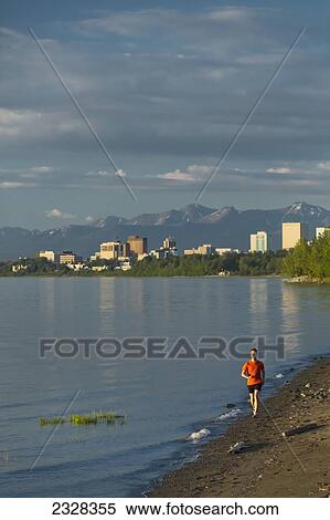 Man Jogging On The Beach Next To The Tony Knowles Coastal Trail During High Tide With The Anchorage City Skyline, Knik Arm, And Chugach Mountains In The Background; Anchorage, Alaska, United States Of America View Large Photo Image Stock Photography - Man Jogging On The Beach Next To The Tony Knowles Coastal Trail During High Tide With The Anchorage City Skyline, Knik Arm, And Chugach Mountains In The Background; Anchorage, Alaska, United States Of America. Fotosearch